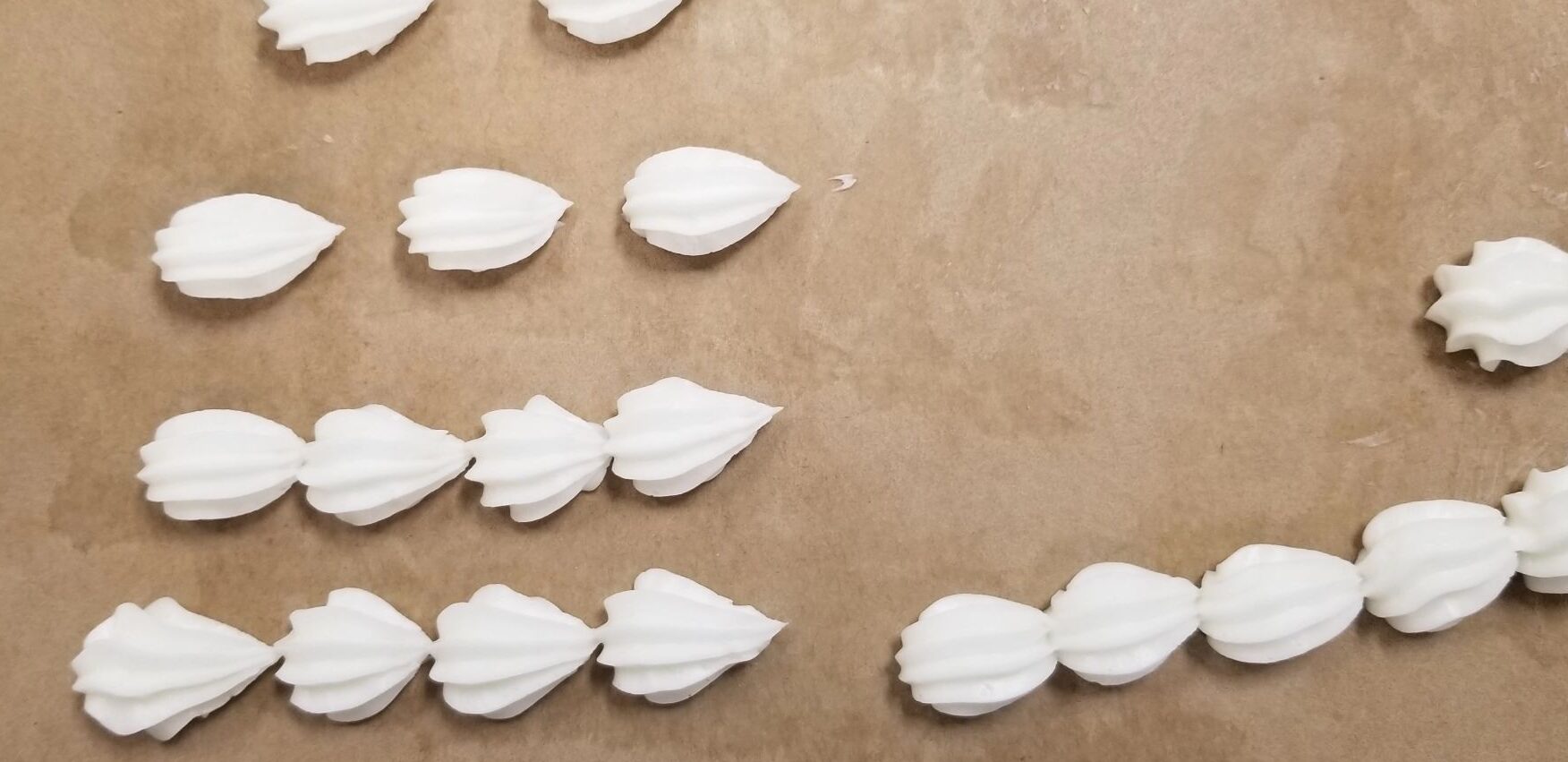 A number of decorative shell shapes piped on brown parchment with white practice "buttercream." Some are by themselves, some are chained together into lines. Some look okay. Many are not quite right. Some are just totally fucked up.