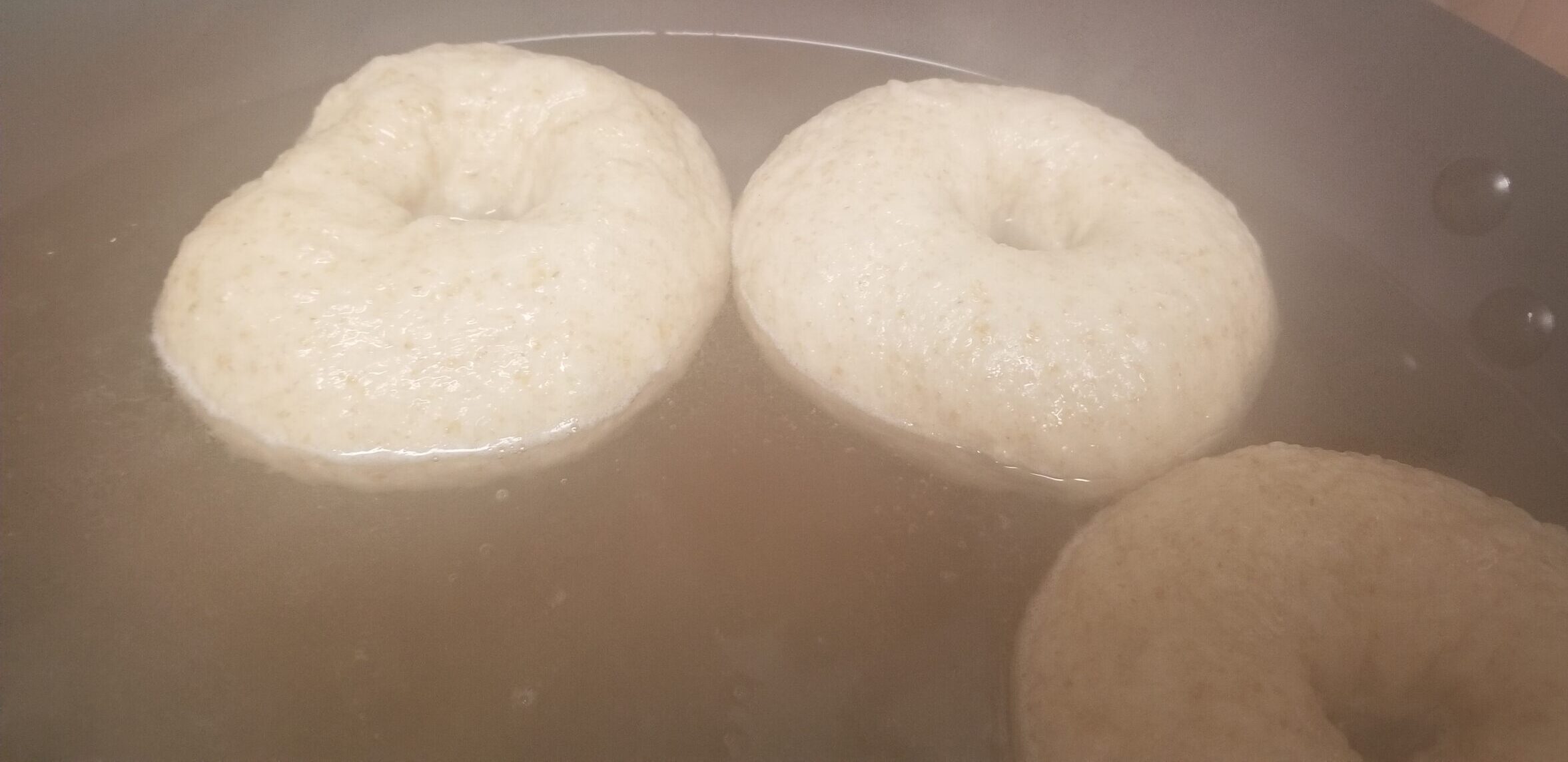 Some unbaked bagels floating in a wok full of boiling water. It's very steamy.
